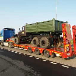 Ciężarówka wywrotka w kolorze khaki transportowana na niskopodwoziowej platformie przez niebieski ciągnik siodłowy, widok z boku na tle asfaltowej drogi i zieleni.