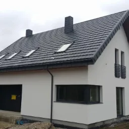 Nowoczesny, parterowy dom z garażem, białą elewacją, grafitową dachówką i oknami dachowymi, na tle lasu i pochmurnego nieba.