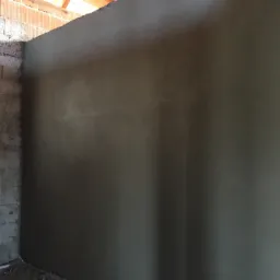 Świeżo nałożony, gładki tynk na ścianie w surowym wnętrzu, widoczne fragmenty cegieł i betonu, przygotowane do dalszych prac wykończeniowych.