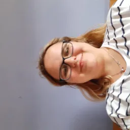 Portret uśmiechniętej blondynki w okularach i koszulce w paski, z delikatnym naszyjnikiem, na tle gładkiej, szarej ściany.