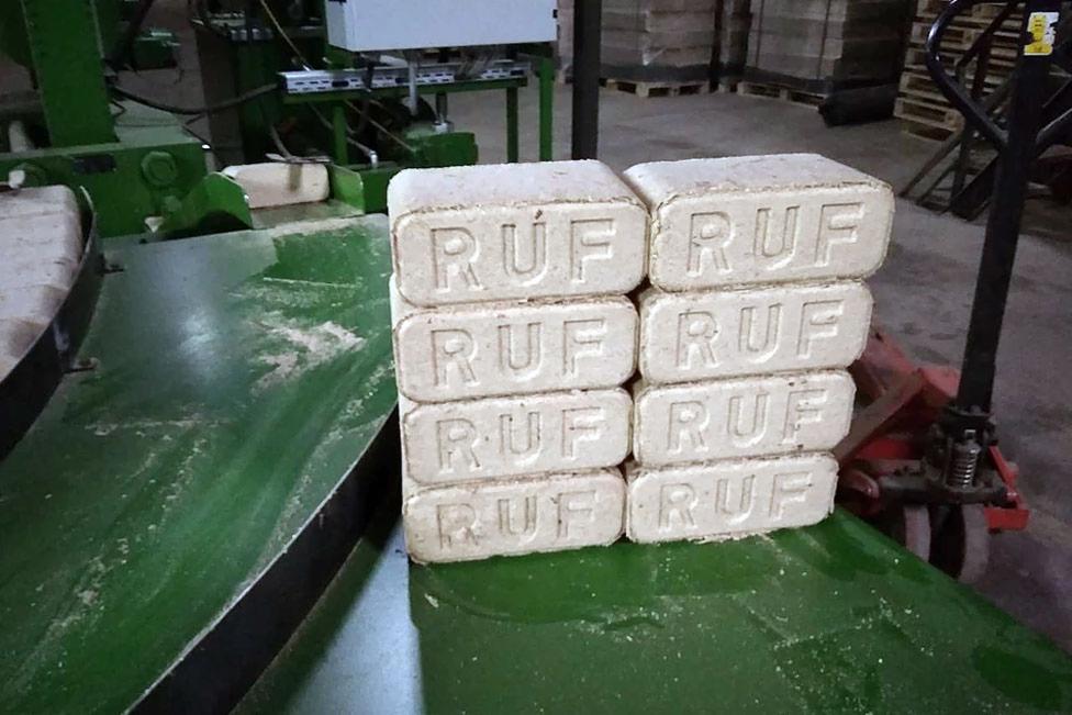 Ułożone w stos brykiety drzewne RUF na zielonej metalowej powierzchni w hali produkcyjnej, z widocznym wózkiem paletowym w tle.