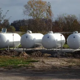 Cztery białe, poziome zbiorniki gazowe ustawione na betonowej podstawie, połączone rurami, na tle jesiennego krajobrazu z drzewami i polami.