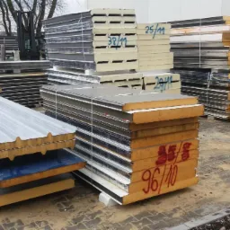 Stosy płyt warstwowych z widocznymi oznaczeniami i numerami na powierzchni, składowane na zewnątrz na placu budowy, w tle widoczny wózek widłowy.