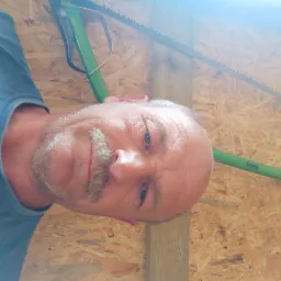 Mężczyzna z piłą kabłąkową na tle drewnianej ściany z desek. Widoczna część twarzy, piła z zieloną rączką i fragment ściany z jasnego drewna.