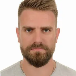 Portret instruktora pierwszej pomocy z brodą, na białym tle, w szarej koszulce. Widoczne zmarszczki mimiczne wokół oczu, co dodaje autentyczności.