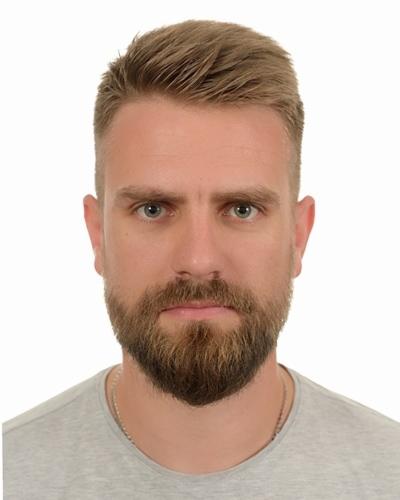 Portret instruktora pierwszej pomocy z brodą, na białym tle, w szarej koszulce. Widoczne zmarszczki mimiczne wokół oczu, co dodaje autentyczności.