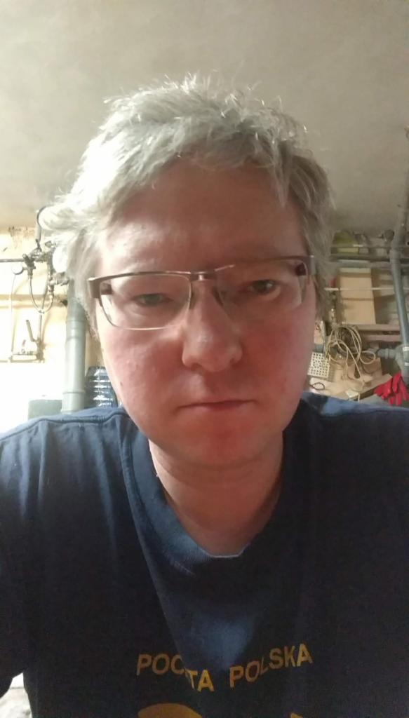 Portret mężczyzny w okularach, koszulka Poczta Polska, instalacje w tle. Ujęcie z dołu, szare włosy, neutralny wyraz twarzy.