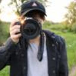 Stowarzyszenie Better Life - Osoba z aparatem fotograficznym skierowanym w stronę obiektywu, na tle zieleni i zachodzącego słońca.