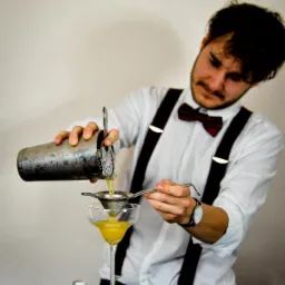 Barman w białej koszuli i z muszką przelewa koktajl z shakera przez sitko do kieliszka martini na czarnym barze.