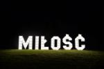 Świecący napis 'MIŁOŚĆ' wykonany z białych, pikselowych liter na ciemnym tle, oświetlony od dołu.