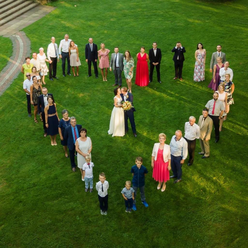 Widok z góry na parę młodą całującą się na środku serca utworzonego przez gości weselnych stojących na trawniku.