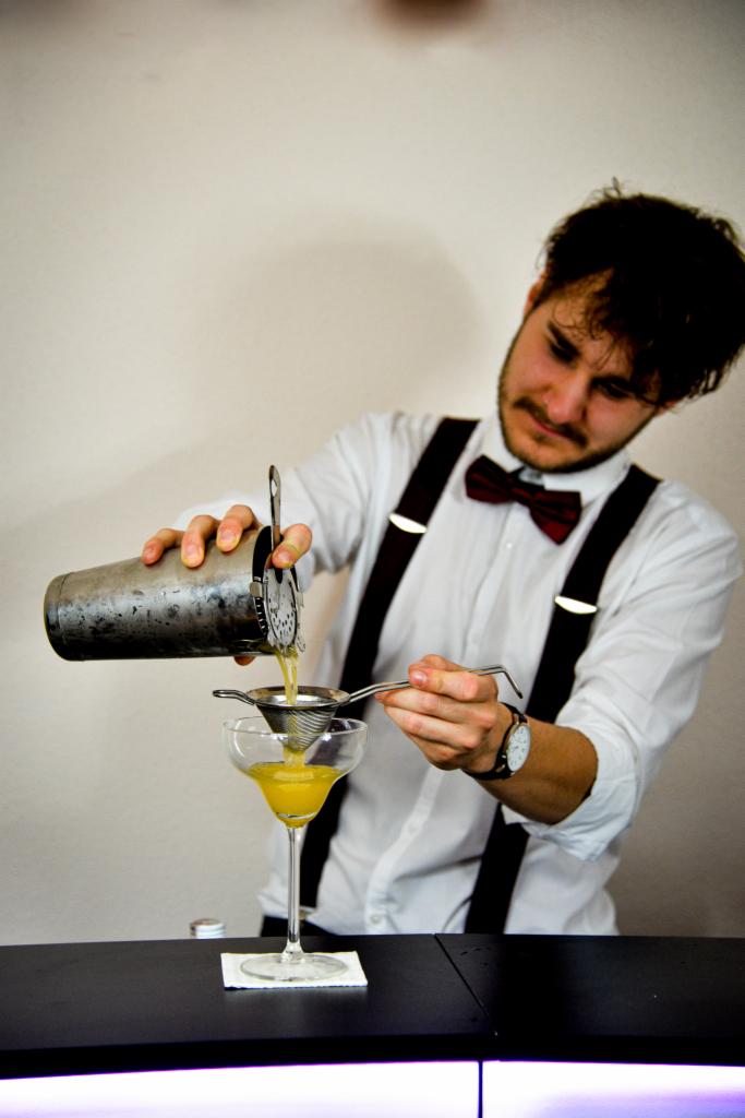 Barman w białej koszuli i z muszką przelewa koktajl z shakera przez sitko do kieliszka martini na czarnym barze.