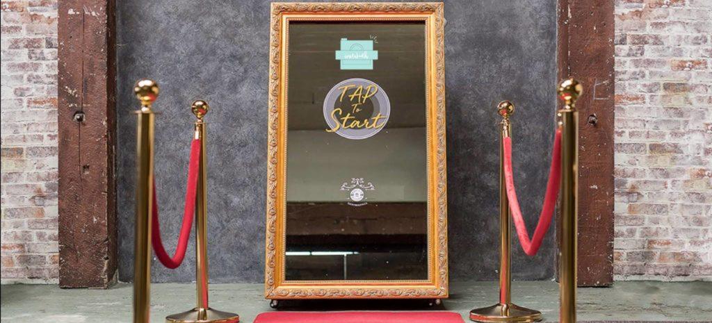 Lustro z napisem 'Tap to Start' w złotej ramie, otoczone złotymi słupkami z czerwonym sznurem, ustawione na czerwonym dywanie na tle ściany z szarego betonu i cegieł.