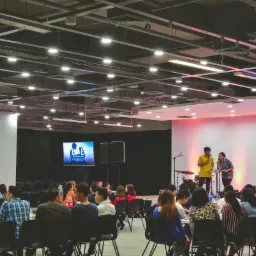 Sala konferencyjna z nagłośnieniem i oświetleniem scenicznym podczas wydarzenia, widoczni prelegenci na scenie i publiczność.