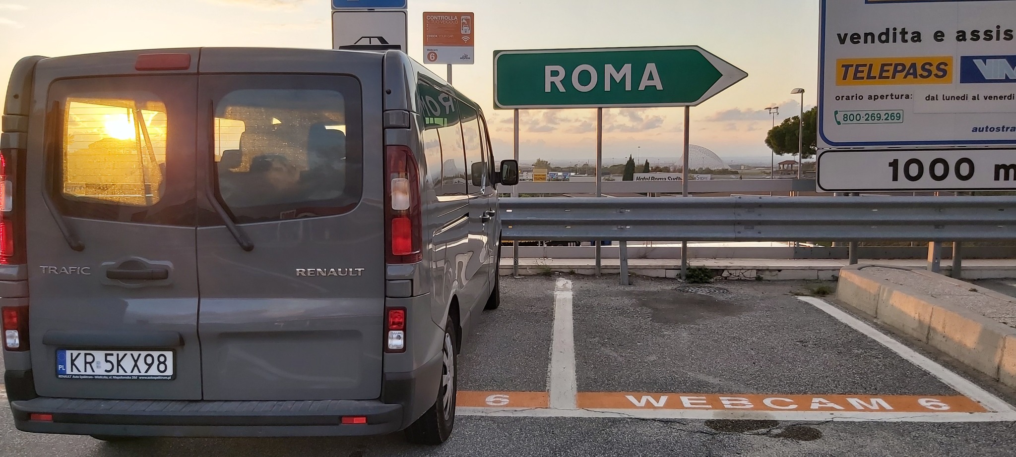 Szary van Renault Trafic z polską rejestracją, zaparkowany na parkingu z oznaczeniem 'WEBCAM 6', znak kierunkowy na Rzym w tle, zachodzące słońce odbija się w tylnej szybie.