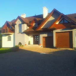 Jasny, dwukondygnacyjny dom z brązowym dachem i drewnianymi elementami wykończeniowymi, widoczny z przodu, w słoneczny dzień. Podjazd wyłożony żwirem.