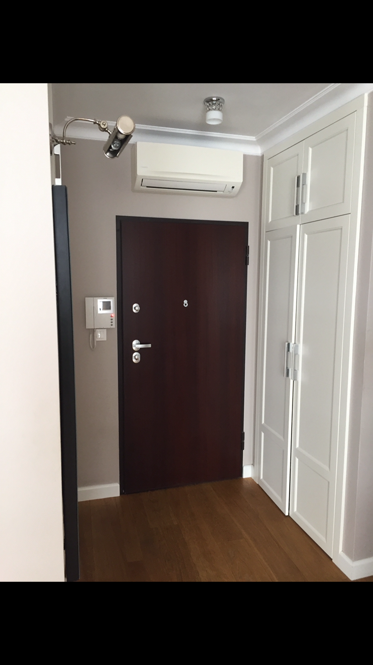 Wnętrze przedpokoju z zainstalowanym klimatyzatorem ściennym nad brązowymi drzwiami wejściowymi z wizjerem, widoczna szafa wnękowa z białymi frontami i drewniana podłoga.