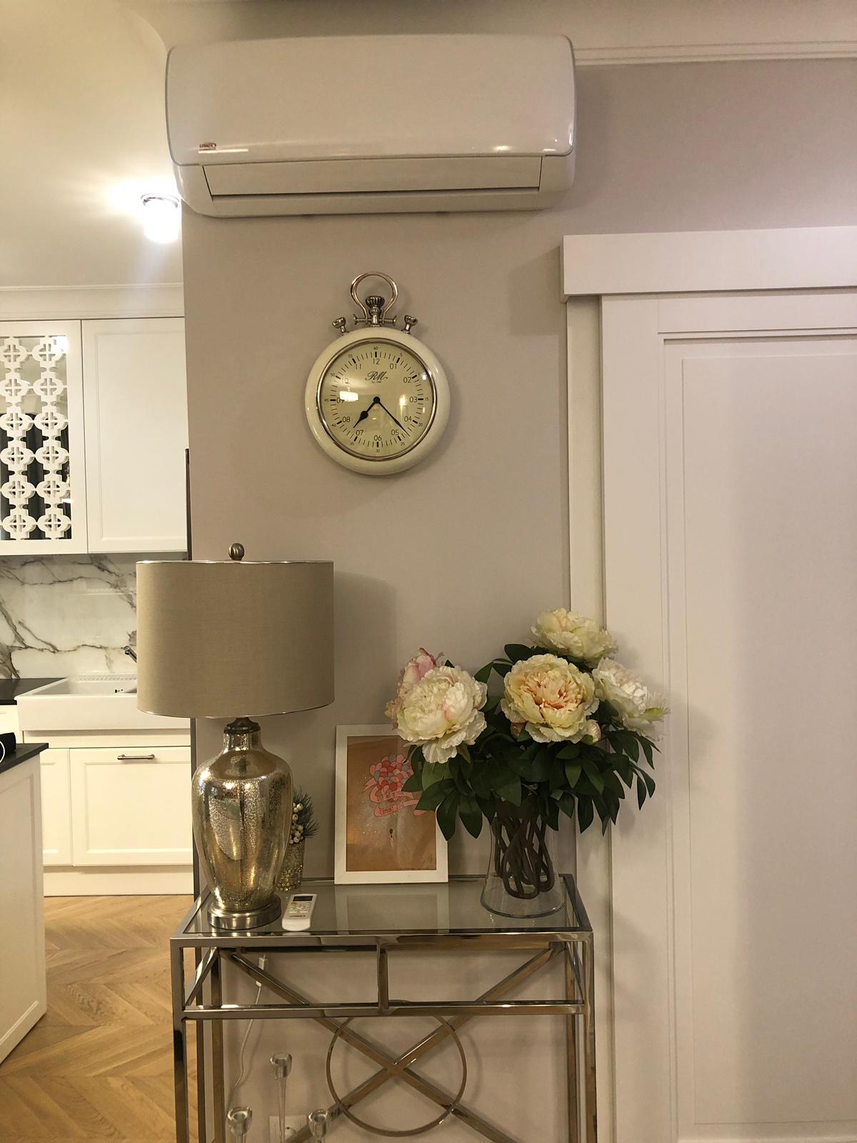 Biały klimatyzator ścienny nad dekoracyjnym zegarem, lampą i bukietem kwiatów w eleganckim wnętrzu.