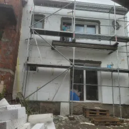 Pracownik na rusztowaniu podczas ocieplania elewacji budynku styropianem, widoczne białe płyty izolacyjne, okna i fragment ceglanego budynku obok.