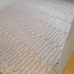 Rozłożona siatka grzewcza Thermoval z czerwonymi kablami na przygotowanym podłożu w rogu pomieszczenia, gotowa do zalania wylewką.