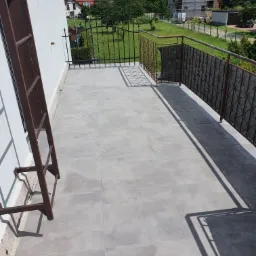 Wyłożony szarymi płytkami balkon z metalową balustradą, widok na okoliczne domy i zieleń, drabina oparta o ścianę budynku.