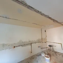 Pokój w trakcie prac przygotowawczych: ściany częściowo pokryte gładzią, widoczne instalacje, drabina z wiertarką, wiadro z zaprawą i narzędzia na podłodze.