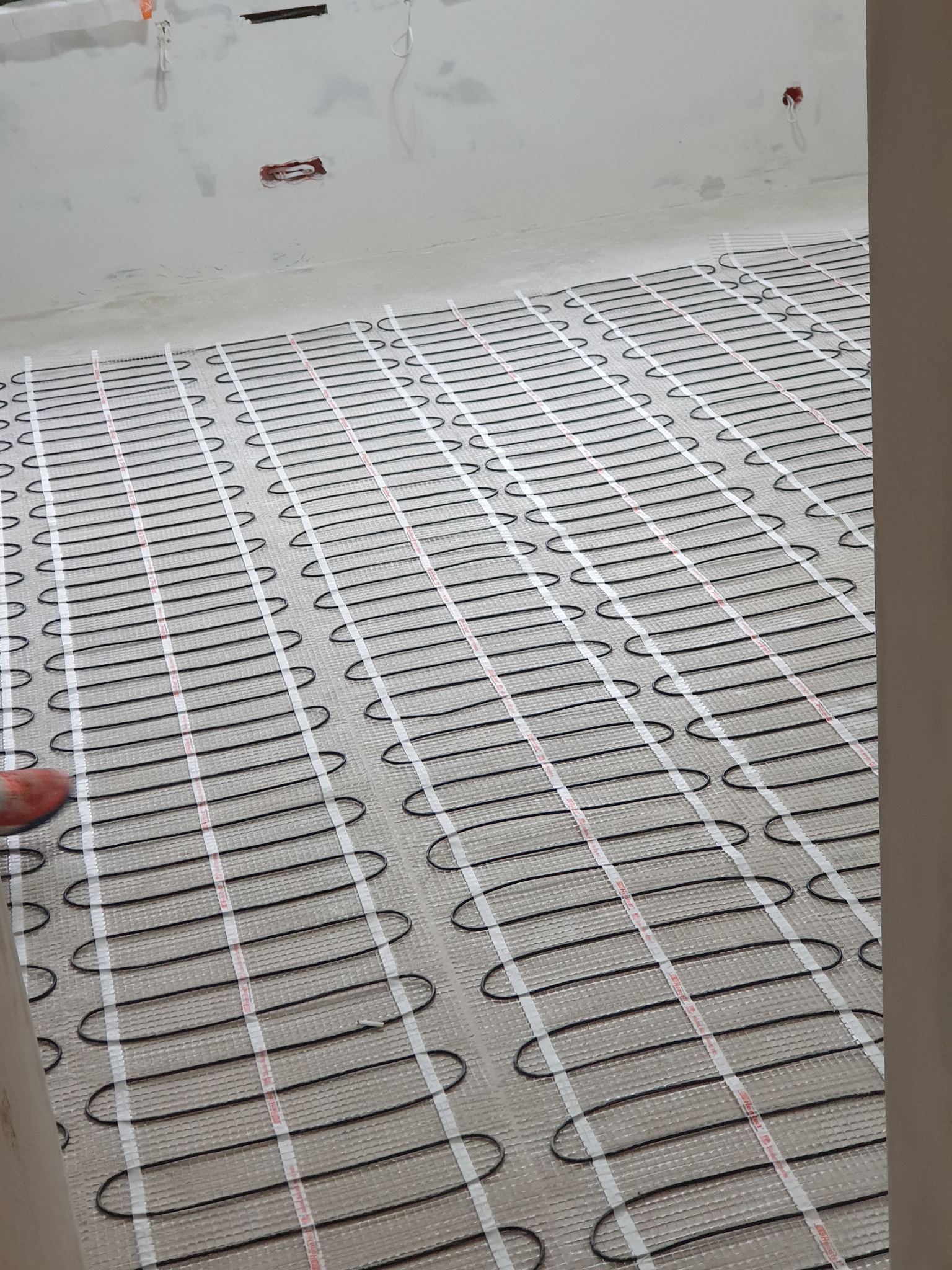 Rozłożona mata grzewcza na podłodze w trakcie montażu, widoczne przewody i taśmy mocujące, surowy stan ścian w tle.