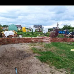 Budowa murku z czerwonej cegły z elementami schodów w ogrodzie, widoczne materiały budowlane i roślinność w tle.