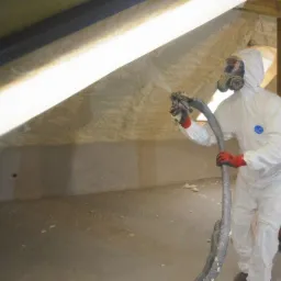 Pracownik w kombinezonie ochronnym i masce natryskuje izolację na poddaszu, widoczna gruba warstwa piany izolacyjnej na skośnej ścianie, oraz fragment konstrukcji drewnianej.