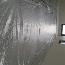 Srebrna folia izolacyjna z widoczną strukturą pęcherzyków powietrza przymocowana do ściany, widoczne zszywki mocujące, w tle okno.
