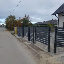Nowoczesne ogrodzenie panelowe z poziomych, grafitowych elementów, osadzone na siatce, widoczne z perspektywy ulicy w jesiennej scenerii.