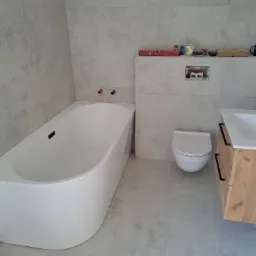 Nowoczesna łazienka z wanną o zaokrąglonych kształtach, podwieszaną toaletą, umywalką z szafką o drewnianym wykończeniu i szarymi płytkami imitującymi beton na ścianach i podłodze. Na ścianie...