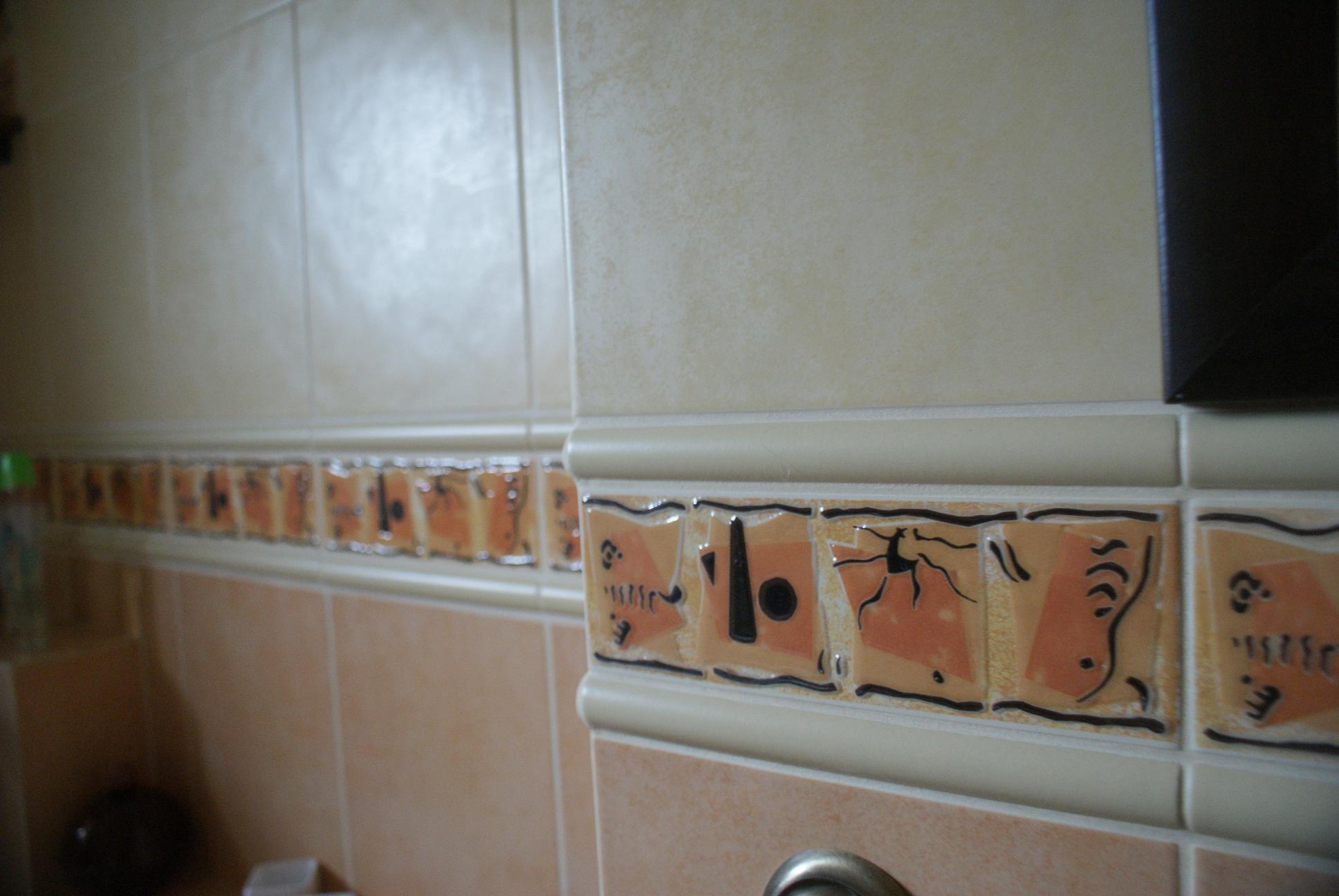 Beżowe płytki ścienne z dekoracyjnym pasem pomarańczowych płytek z czarnymi wzorami, widoczne fugi i fragment baterii.