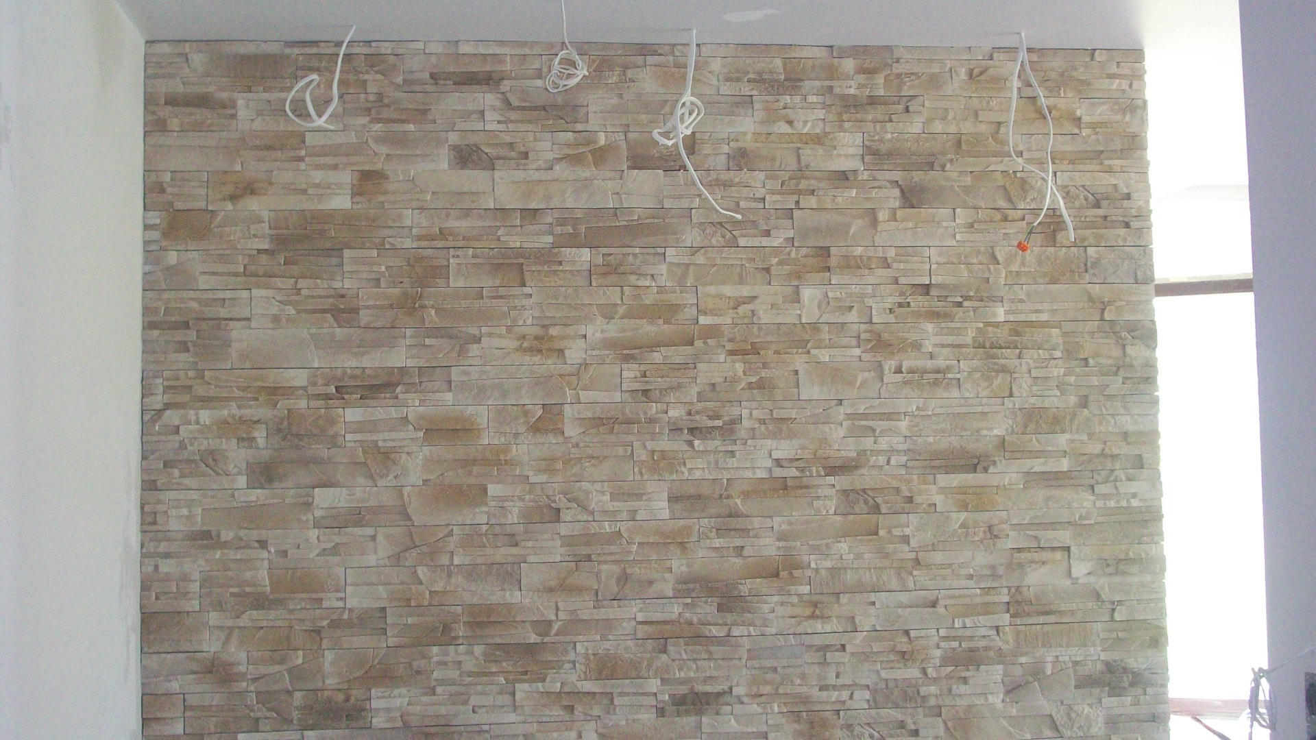 Ściana wyłożona dekoracyjnym kamieniem w odcieniach beżu i brązu, widoczne niedokończone podłączenia elektryczne wystające z sufitu, wnętrze w trakcie wykańczania.
