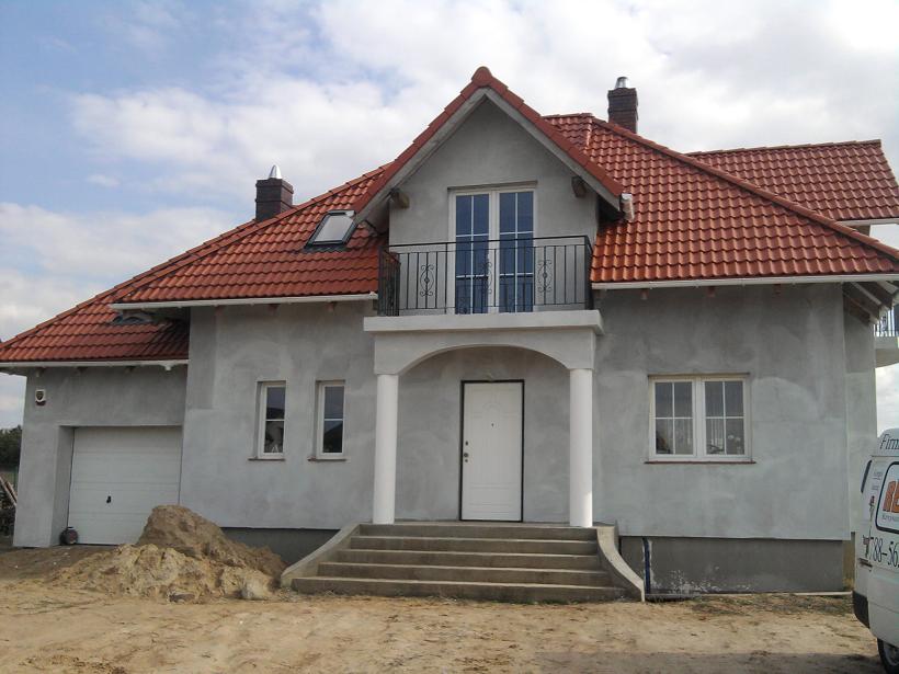 Nowo wybudowany dom jednorodzinny z czerwoną dachówką i elewacją w trakcie wykończenia, widoczny stos piasku przed garażem.