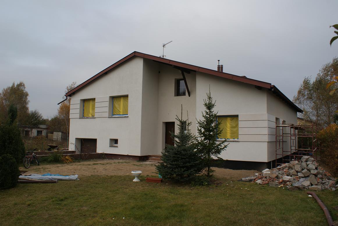 Biała elewacja domu jednorodzinnego w trakcie prac, okna zabezpieczone żółtą folią, widoczne rusztowanie i gruz na trawniku, w tle budynki gospodarcze.