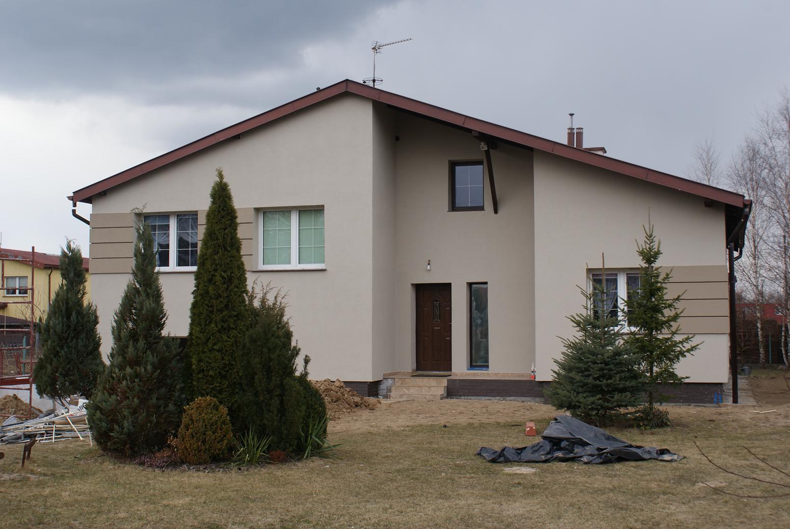 Dwukondygnacyjny dom z beżową elewacją, brązowym dachem i stolarką okienną, widoczny z przodu, z anteną na dachu i iglakami w ogrodzie, na działce w trakcie prac ziemnych.