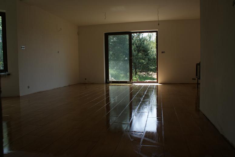 Świeżo położona, błyszcząca podłoga z płytek imitujących drewno w pustym pokoju w Osielsku, z widokiem na zieleń za oknem.