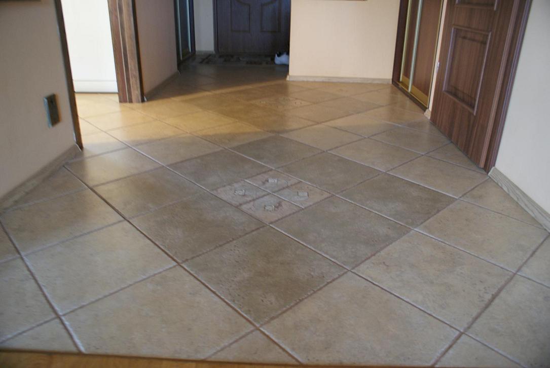 Beżowa podłoga wyłożona kwadratowymi płytkami ułożonymi romboidalnie, z dekoracyjnym motywem na środku, widoczna w korytarzu z drewnianymi drzwiami i ościeżnicami.