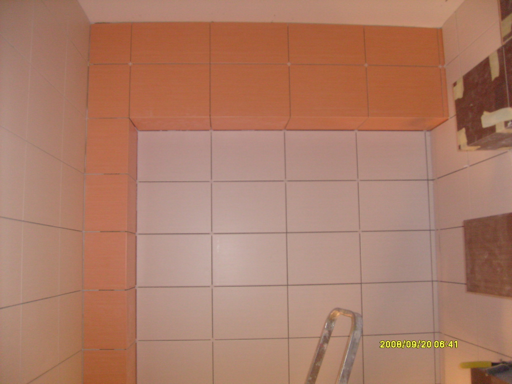 Wykończona ściana w łazience z białymi i pomarańczowymi kwadratowymi płytkami, widoczny fragment drabiny malarskiej.