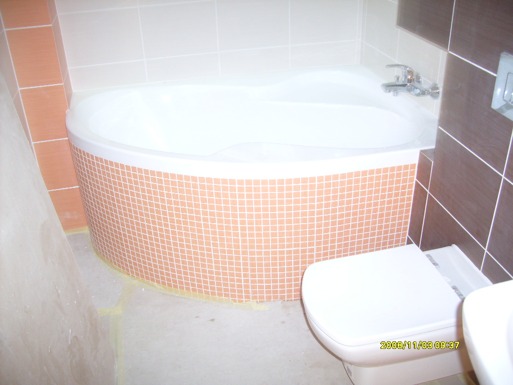 Narożna wanna obudowana mozaiką w odcieniach brzoskwini, widoczna również toaleta i fragment umywalki, w tle brązowe i białe płytki ścienne, widoczne ślady po remoncie na podłodze.