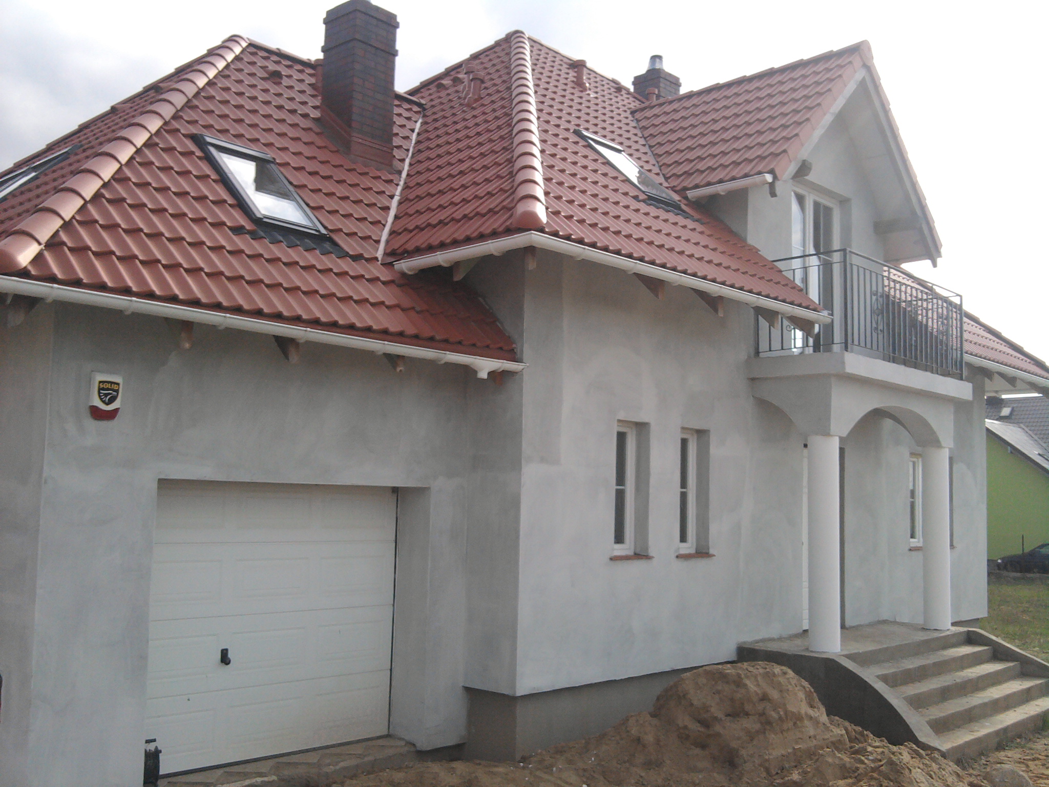 Dwupiętrowy dom z garażem w stanie surowym, z czerwoną dachówką, widok z zewnątrz, przed domem hałda ziemi.