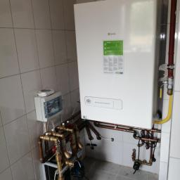 Sebterm Technika Grzewcza i Klimatyzacyjna Sebastian Kramarz - Nowy, biały kocioł gazowy marki Beretta, model Mynute Boiler Green, zamontowany na ścianie z jasnych, kwadratowych płytek, z widocznymi rurami miedzianymi i zaworami, podłączony do instalacji,...