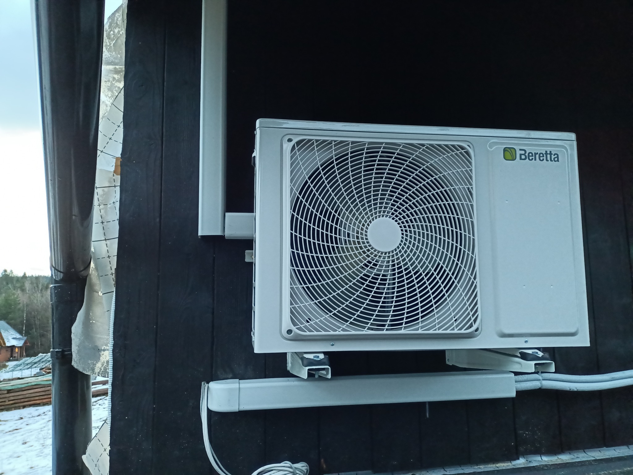 Zewnętrzna jednostka klimatyzacji marki Beretta zamontowana na ciemnej, drewnianej ścianie budynku. Widoczne przewody i elementy montażowe.