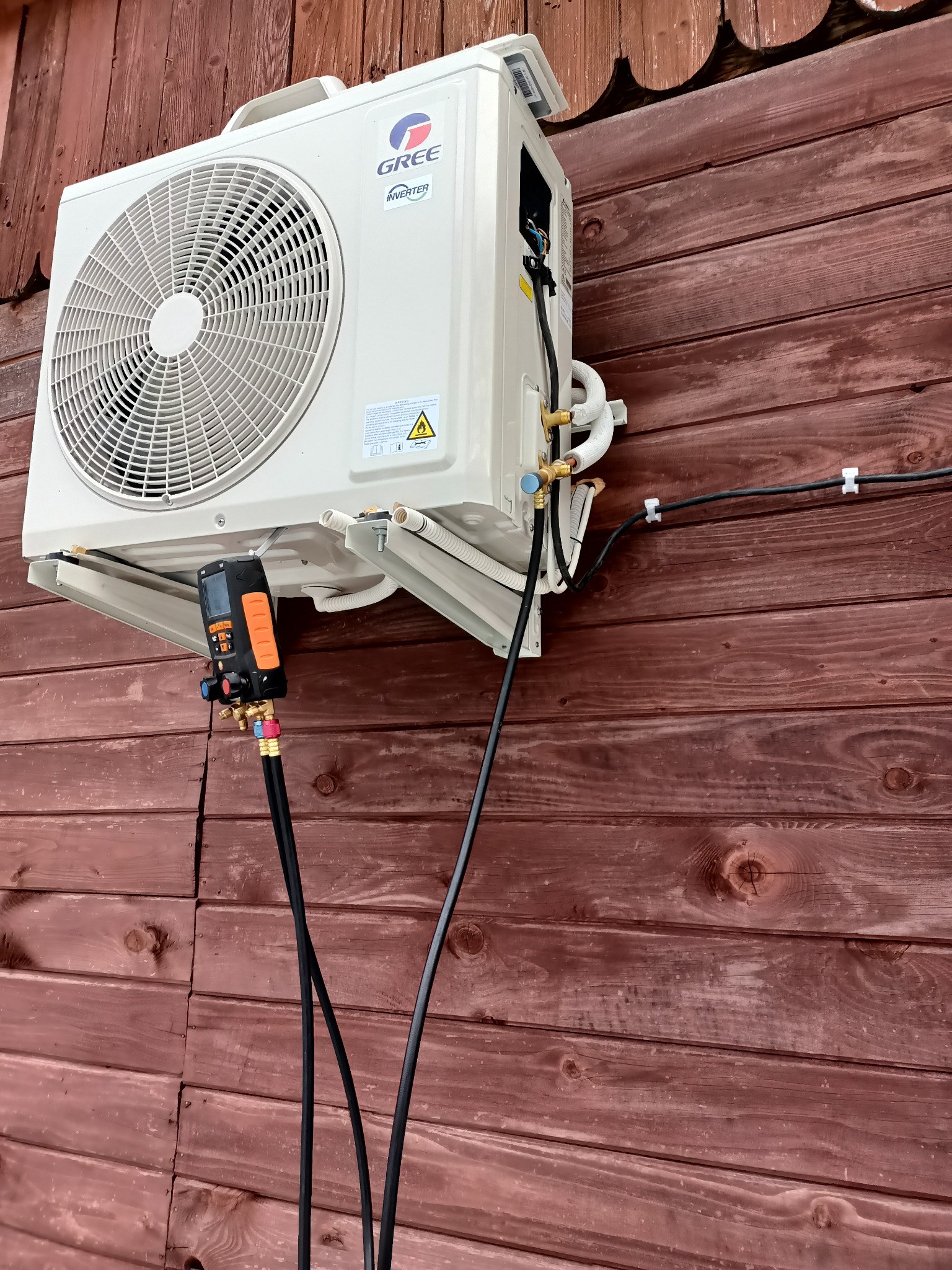 Biała jednostka zewnętrzna klimatyzacji marki Gree zamontowana na brązowej drewnianej ścianie, podłączona do pompy próżniowej za pomocą węży, widoczne przewody elektryczne i rurki chłodnicze.