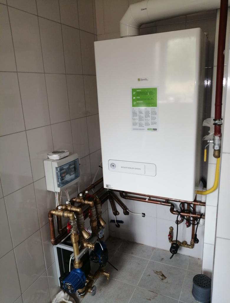 Nowy, biały kocioł gazowy marki Beretta, model Mynute Boiler Green, zamontowany na ścianie z jasnych, kwadratowych płytek, z widocznymi rurami miedzianymi i zaworami, podłączony do instalacji,...
