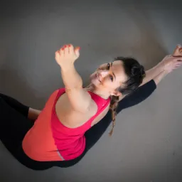 Kobieta w czarnych legginsach i czerwono-pomarańczowym topie wykonuje pozycję jogi, siedząc na szarym tle i uśmiechając się w górę.