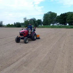 Czerwony traktor z operatorem przygotowuje pole pod zasiew, widoczne ślady opon i spulchniona gleba, w tle zieleń drzew.