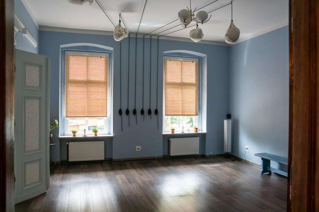 Jasne, przestronne wnętrze z drewnianą podłogą, błękitnymi ścianami, dwoma oknami z bambusowymi roletami i nietypowym oświetleniem sufitowym z elementami przypominającymi worki.