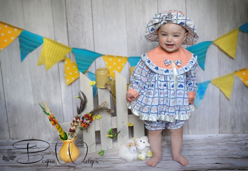 Uśmiechnięta dziewczynka w kwiecistym stroju i kapeluszu, pozująca na tle drewnianej ścianki z girlandami, obok wazon z palmą wielkanocną i pluszowe owieczki.